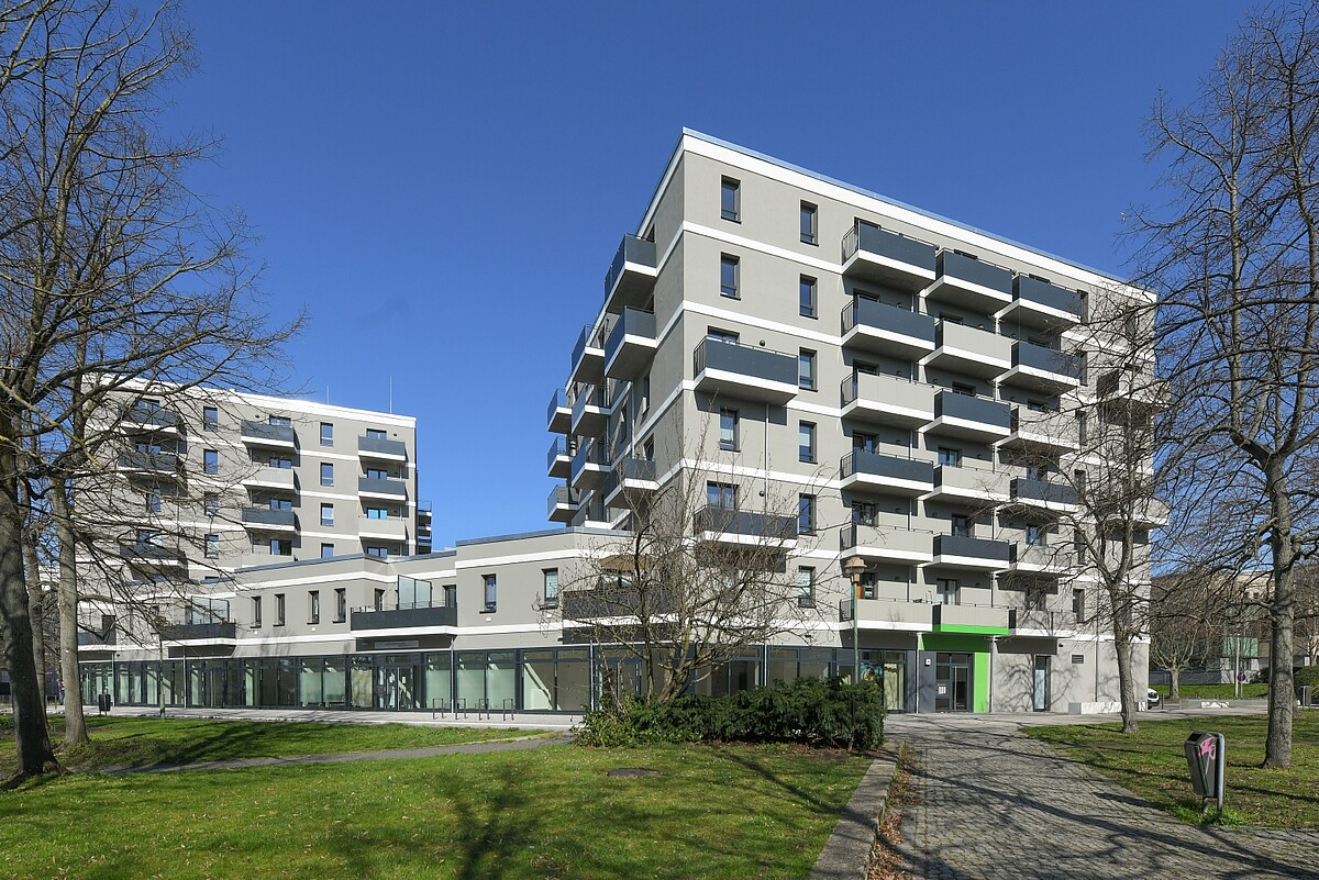 Das Foto zeigt einen Neubaukomplex. Dieser ist mehrgeschossig. Einige Wohnungen verfügen über Balkone. Die Fassade ist in einem Grau gestrichen. Vor dem Neubau befindet sich eine grüne Wiese. Darauf stehen Bäume. Es ist Frühling.