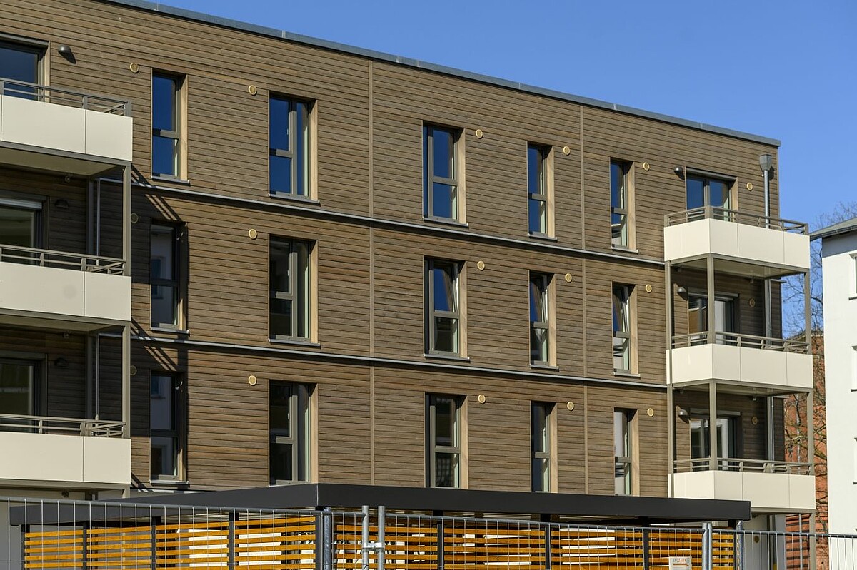 Auf dem Bild ist die Nahaufnahme einer Neubaufassade zu sehen. Diese besteht zu großen Teilen aus Holz. Dieses ist dunkelbraun lackiert. Der Neubau hat zudem 6 Balkone.