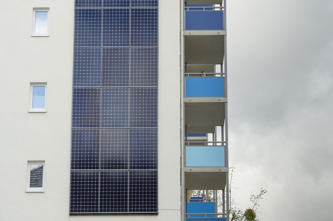 An der Fassade eines Gebäudes befindet sich eine Photovoltaikanlage. Von dieser ist eine Nahaufnahme gemacht worden. Im Hintergrund sind Balkone zu sehen. Der Himmel ist bewölkt.